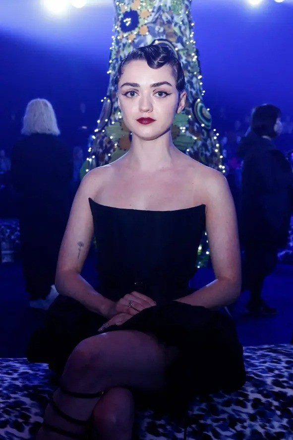 Diễn viên Maisie Williams ngồi trong không gian show diễn như một hòn đảo ở xứ sở thần tiên, nơi ánh sáng xanh và họa tiết hoa sặc sỡ bao trùm.
