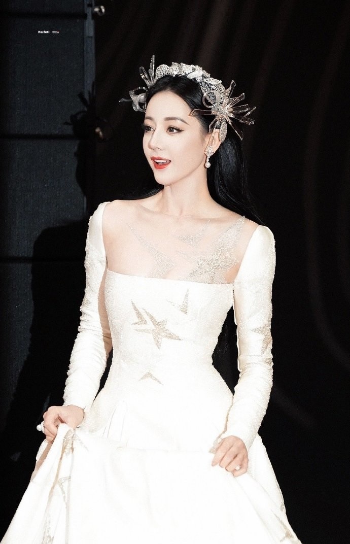 Địch Lệ Nhiệt Ba mặc đầm trắng cổ vuông, màu trắng ánh sao, trang điểm nổi bật. Cô đội kèm mấn đính hoa và cườm.