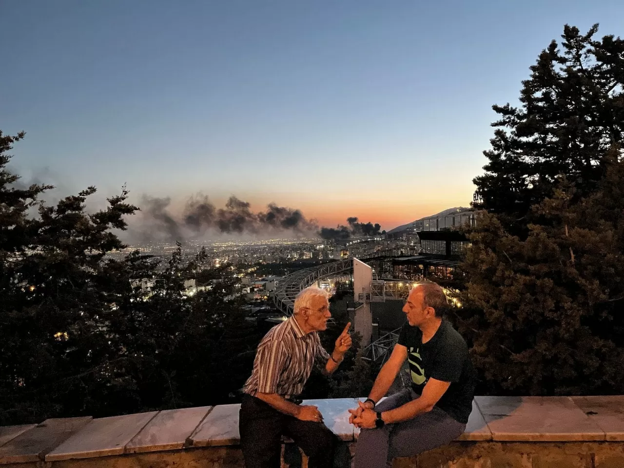 Hai người đàn ông ở Tehran, Iran, tranh luận về xung đột giữa Iran và Israel vào thứ Ba, ngày 17 tháng 6, trong khi phía sau họ là khói bốc lên từ một nhà máy lọc dầu đang cháy sau khi bị Israel tấn công. (Nguồn: Redux)