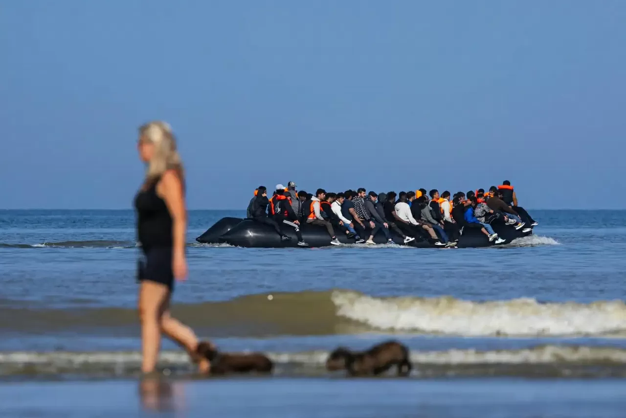 Một nữ du khách dắt chó đi dạo trong lúc phía sau, một nhóm người đang lên thuyền buôn lậu để vượt eo biển Manche, bãi biển Hardelot, Pháp. Trong vòng vài giờ ngày 30/6, năm chiếc thuyền chở hàng chục người đã rời khỏi các bãi biển Hardelot và Equihen trong cảnh hỗn loạn, giữa lúc cảnh sát Pháp can thiệp bằng hơi cay và lực lượng cứu hộ túc trực gần bờ. Một số người phải lội nước đến tận cổ để tiếp cận các thuyền phao, trong khi trẻ nhỏ được cõng trên vai. Riêng trong ngày này, hơn 879 người di cư đã đến được bờ biển Anh, con số cao thứ ba từ đầu năm. (Nguồn: Getty Images)