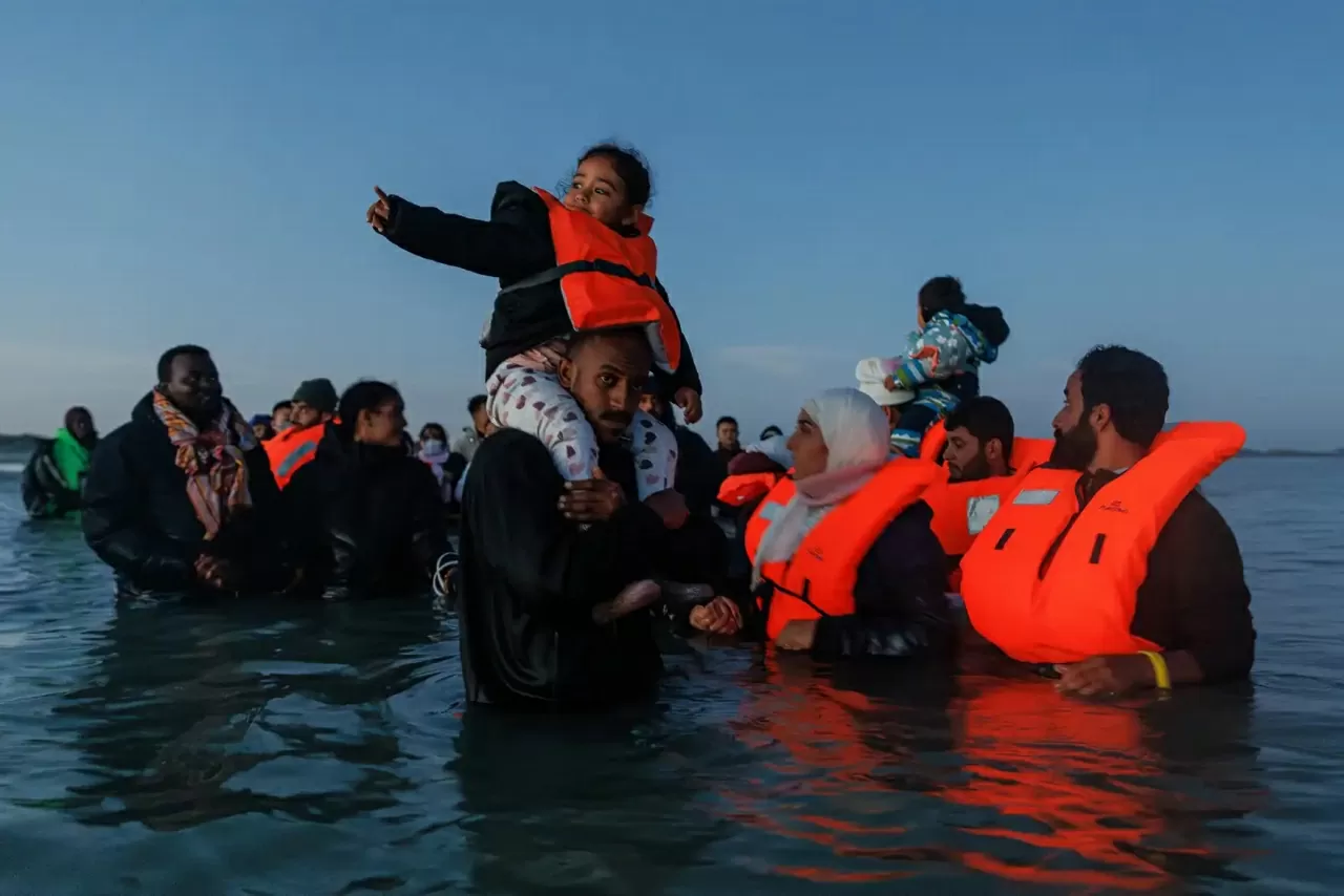 Một người cha bế con gái nhỏ trong khi nhiều người khác chờ đợi dưới làn nước ngoài khơi Gravelines (Pháp) để lên thuyền vượt eo biển Manche sang Anh. Bộ Nội vụ Anh xác nhận người di cư đầu tiên đã bị trục xuất về Pháp theo thỏa thuận gây tranh cãi “một v